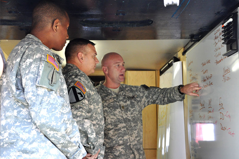 A major pointing to procedures written on a whiteboard as a colonel and command sergeant major look on