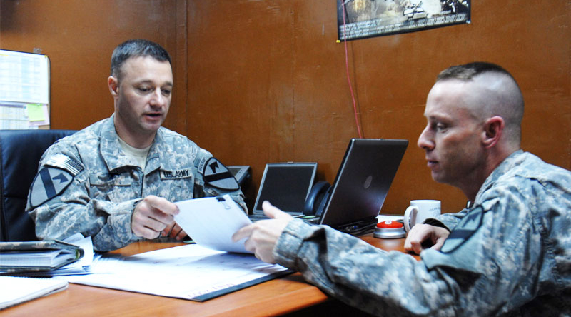 One Soldier delivering feedback to another Soldier at a desk in an office